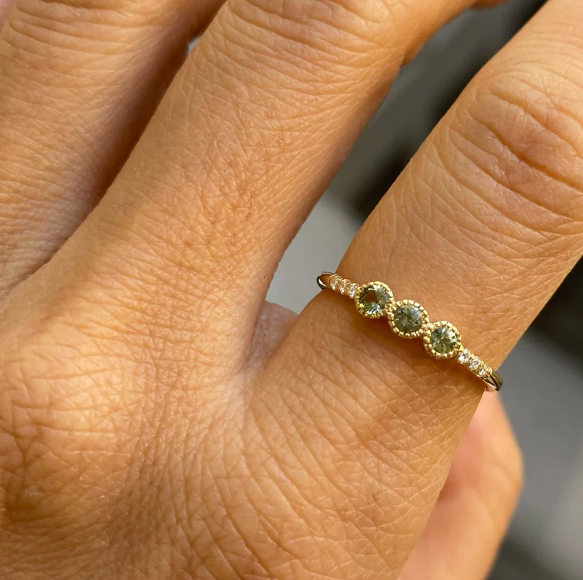 3 light green sapphires set in milgrain detailed bezel settings in a row at the center of a thin gold band. To add even more texture, there are three additional smaller white diamonds flush set into the band on either side of the large central diamonds. The slight texture makes this otherwise clean design feel ever so intimate on model