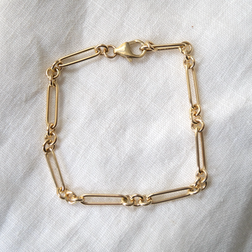 Gold bracelet with mixed link designs, including elongated and rounded links, displayed on a white textured fabric.