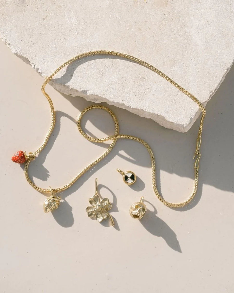 A gold necklace with a lobster clasp, featuring Satomi Studio's Skull Charm pendants, creatively displayed on a textured white stone against a beige surface.