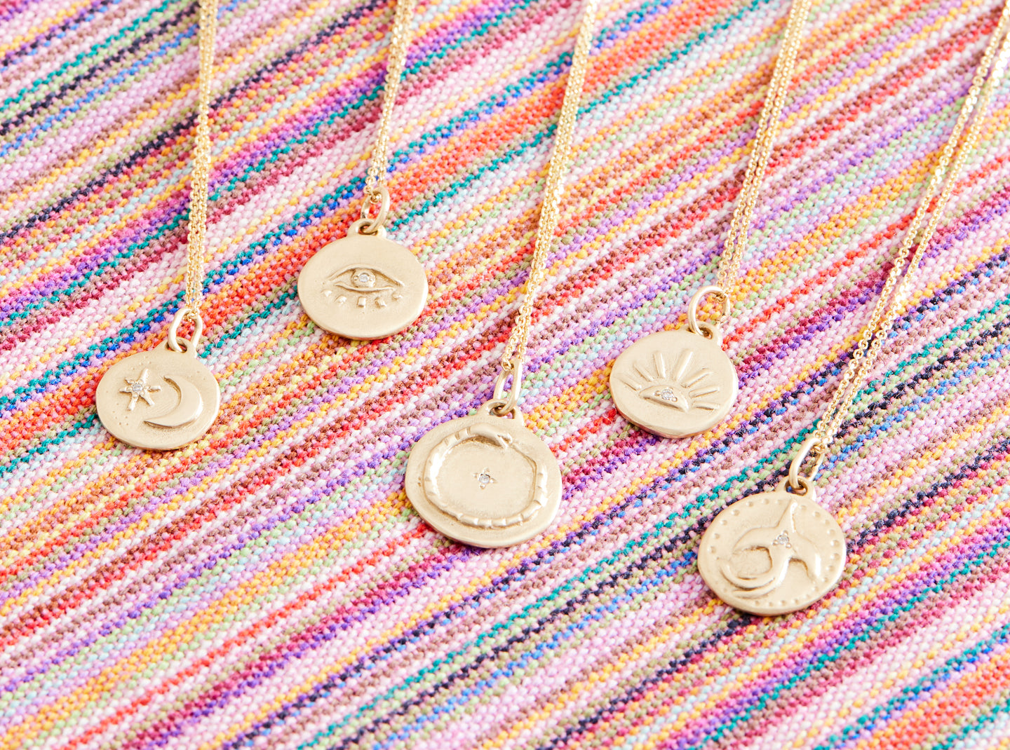 Five gold necklaces on gold chain, each one with a different symbol: moon with star, eye with lashes, snake eating its tail, rising sun with rays, bird taking flight. All of them have diamonds set in the center