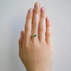 2 of 3: A vibrant emerald set in a bezel setting with engraved lines, flanked by two round diamonds and pave diamonds on either side, on hand