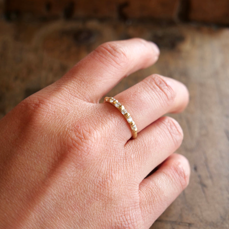 A close-up image of a hand wearing a delicate 18k gold ring set with multiple small Cora Classic - Antique Diamonds, against a wooden surface background by Marisa Mason Jewelry.