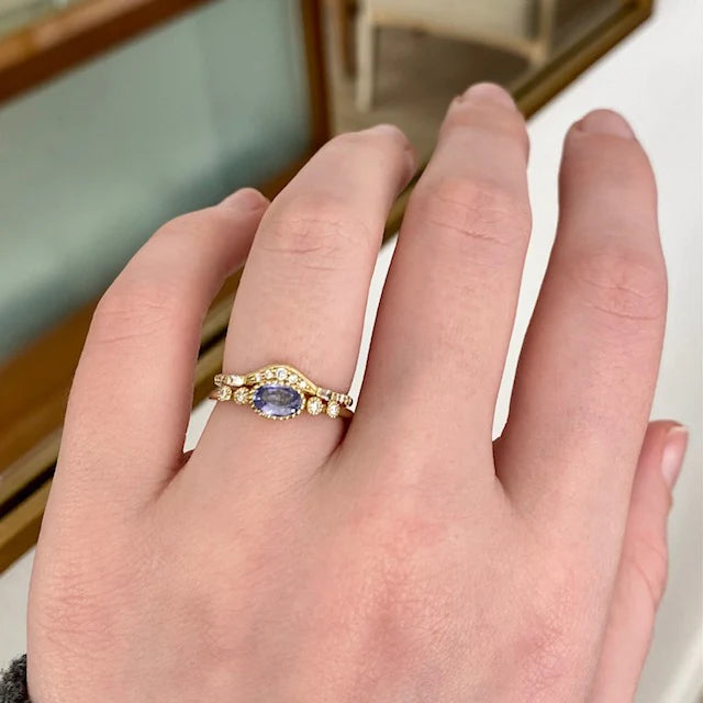 A thin gold band with a bezel set oval blue sapphire in the center, and two round white diamonds on either side, on hand