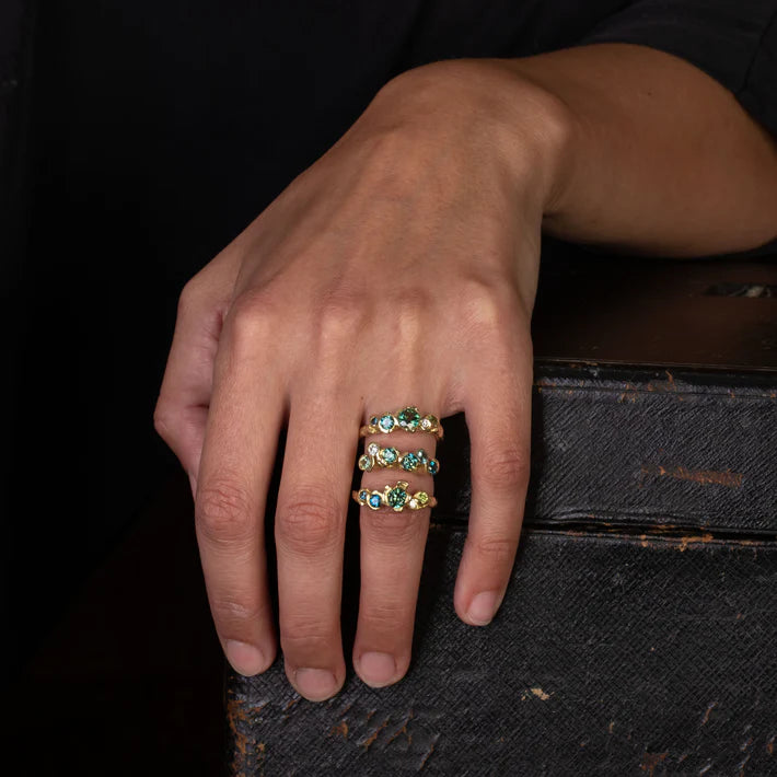 Organically shaped gold band set with four green and blue sapphires, and one white diamond on model with other rings