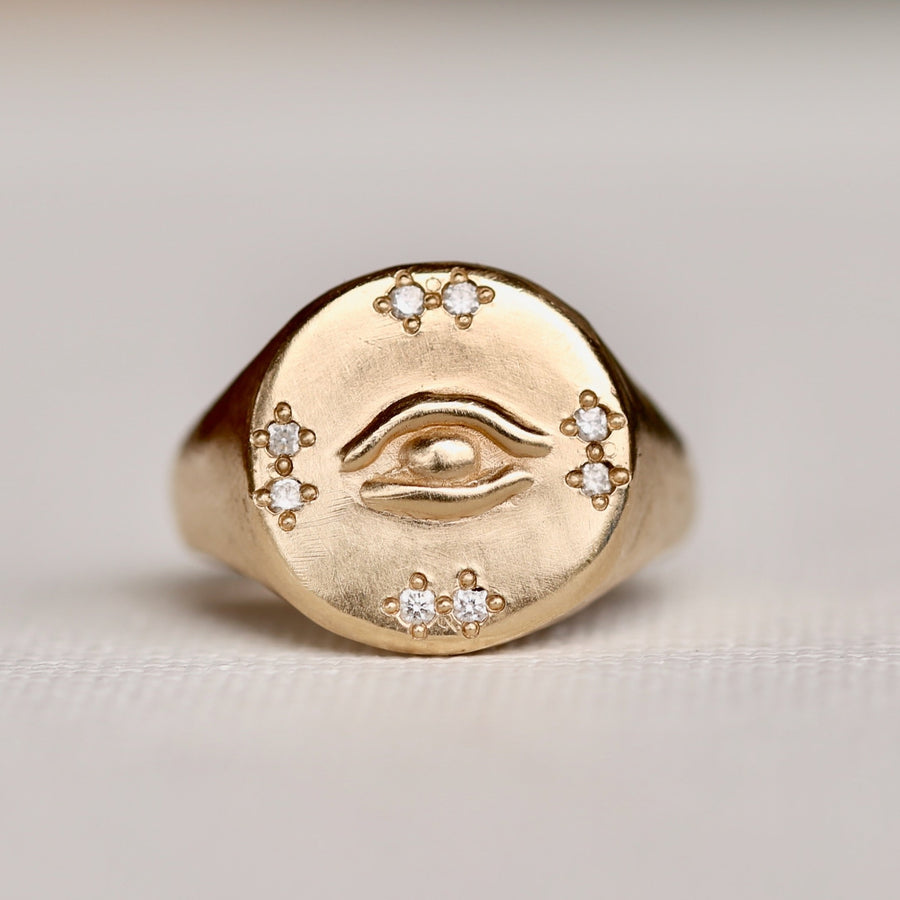 A round 14k signet ring with a hand carved eye at the center, and 6 bead set diamonds set above, below and on either side of the eye