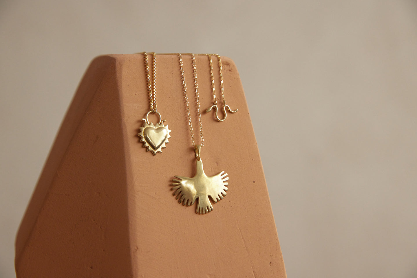 Three brass necklaces with a heart, a snake, and a bird charm hang on pink wood block.Brass and Sterling Silver necklaces, delicate and statement necklaces, evil eye charm, delicate charms, 14k gold necklaces, brass necklaces, handmade jewelry in Oakland.