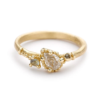 A pretty solitaire style engagement ring with a pear shaped pale champagne diamond flanked by a grey diamond and a green sapphire, and surrounded by delicate gold work on a matte band.
