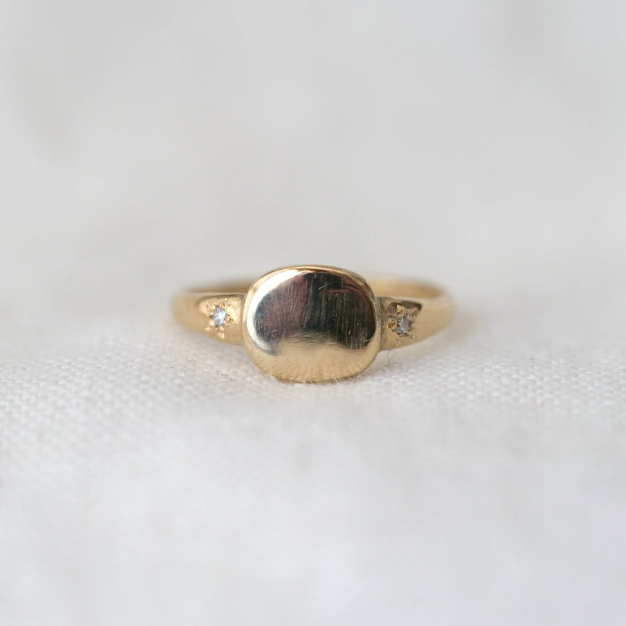 Gold signet ring with a circular design and two star set diamonds on either side, on a white background