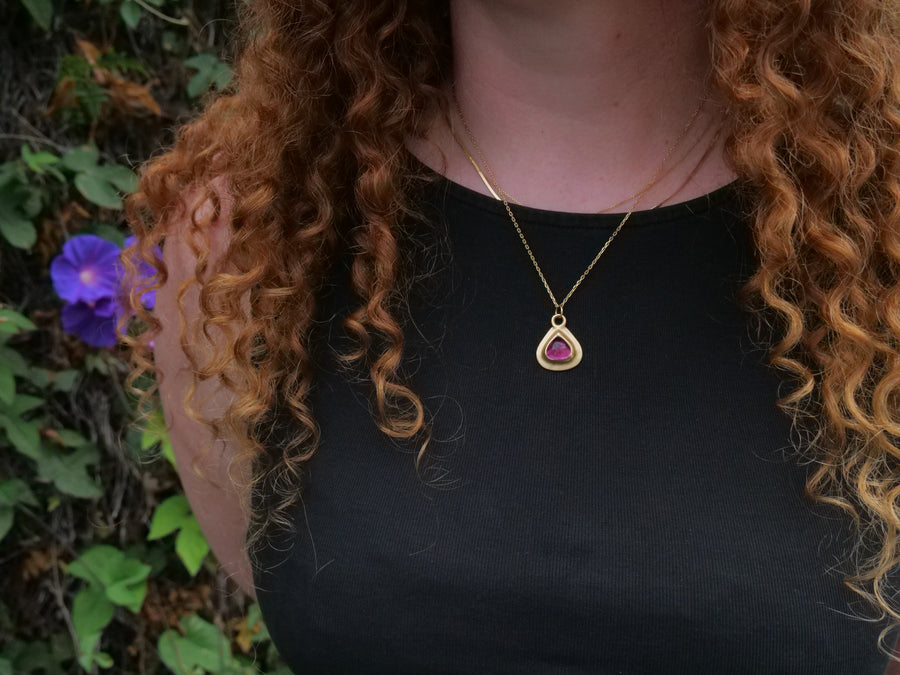 Person wearing a gold necklace with a pink pendant against a natural background