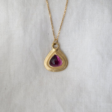 Gold necklace with a gold teardrop-shaped pendant featuring a purple gemstone on a light gray background