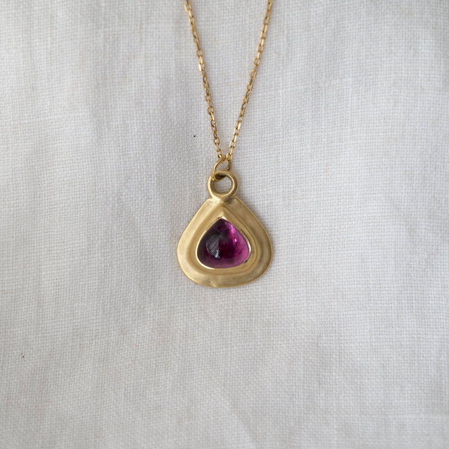Gold necklace with a gold teardrop-shaped pendant featuring a purple gemstone on a light gray background