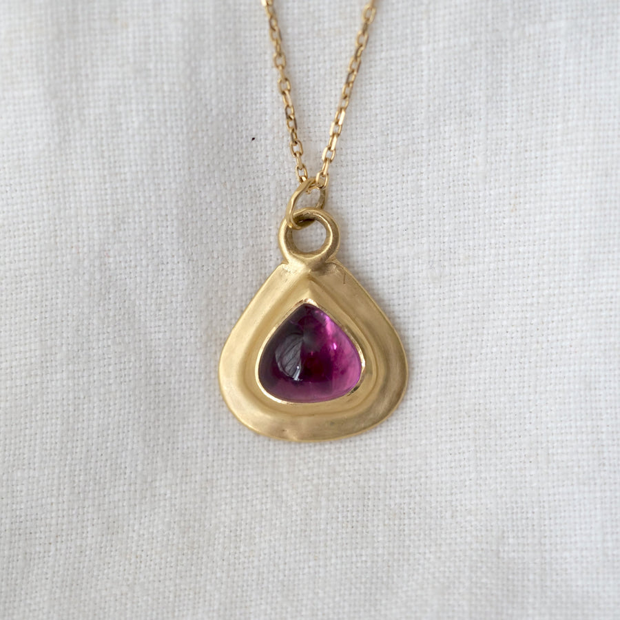 Gold necklace with a teardrop-shaped pendant featuring a purple gemstone on a light gray background