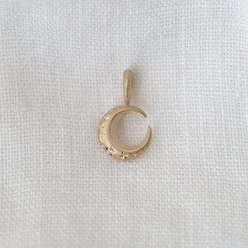 Gold crescent moon pendant with engraved designs of stars and a moon, with two white diamonds, on a light fabric background