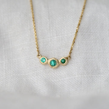 Gold necklace with three bezel set emerald stones on a white fabric background