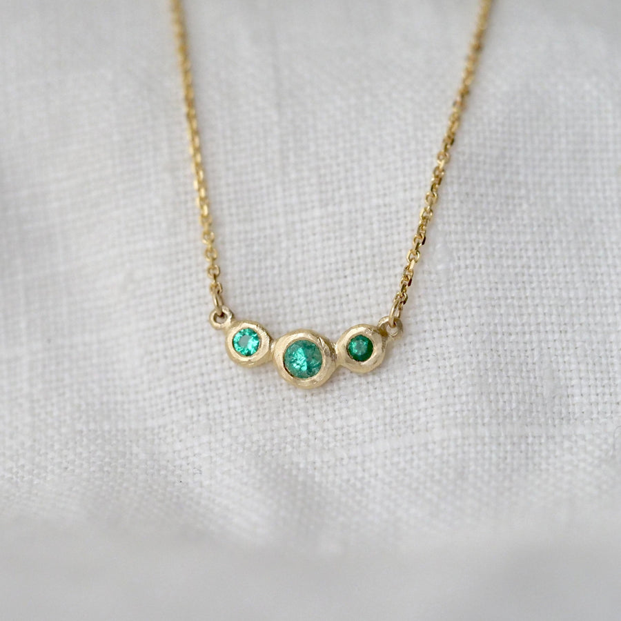 Gold necklace with three bezel set emerald stones on a white fabric background