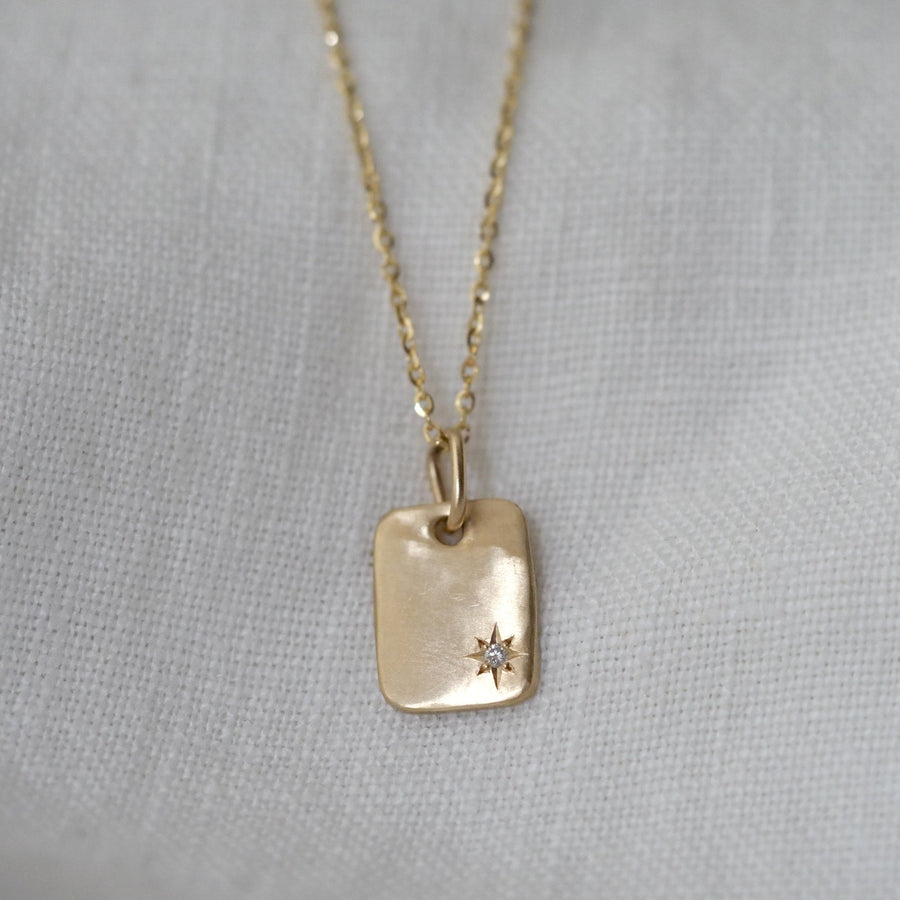 Gold chain with a rectangular dog tag pendant with a single star set diamond on a light gray background