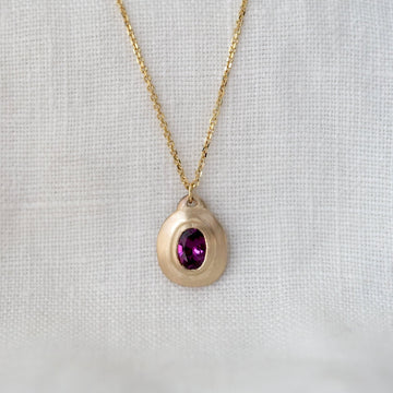 Gold necklace with a teardrop-shaped pendant featuring a purple gemstone on a light gray background