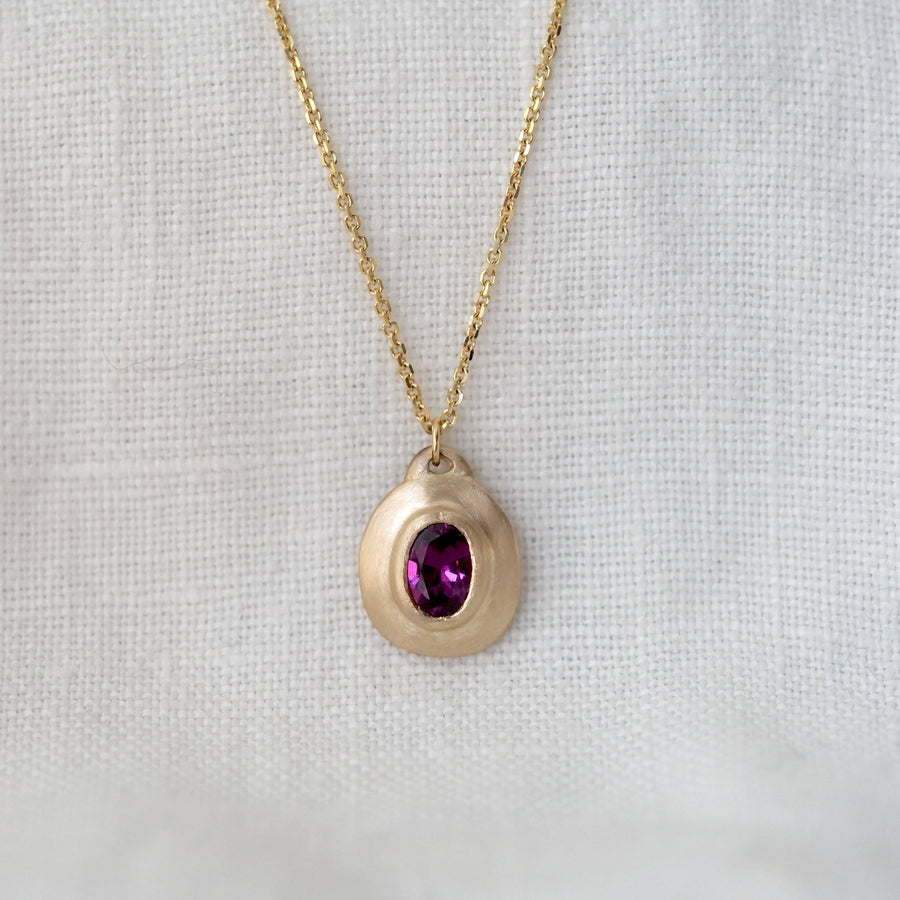 Gold necklace with a teardrop-shaped pendant featuring a purple gemstone on a light gray background