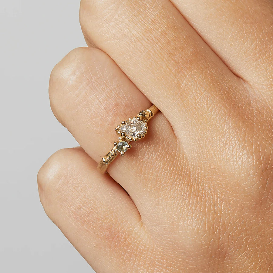 A pretty solitaire style engagement ring with a pear shaped pale champagne diamond flanked by a grey diamond and a green sapphire, and surrounded by delicate gold work on a matte band, on models hand