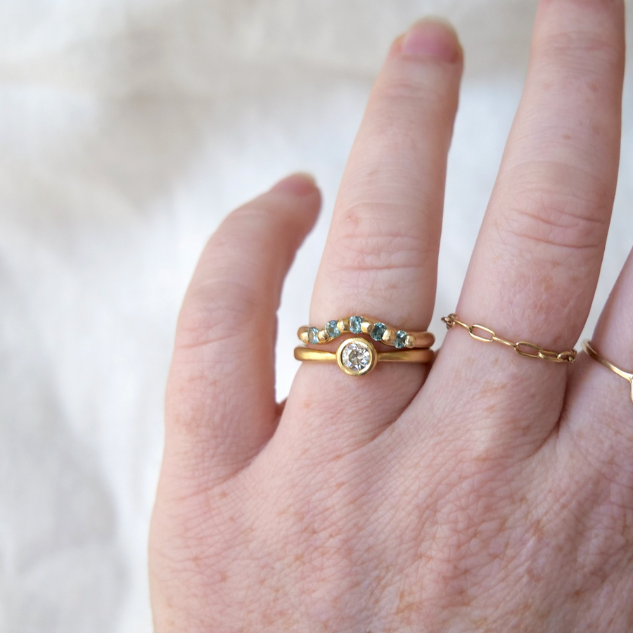 Rounded gold band with carved gold granulation details used to set five montana sapphires stones, creating a curve in the band that makes room for wearing with a solitaire ring on model