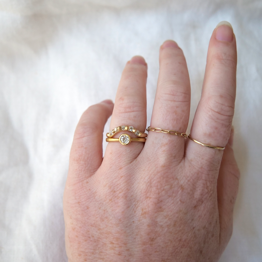 Rounded gold band with carved gold granulation details used to set five diamonds stones, creating a curve in the band that makes room for wearing with a solitaire ring shown on model