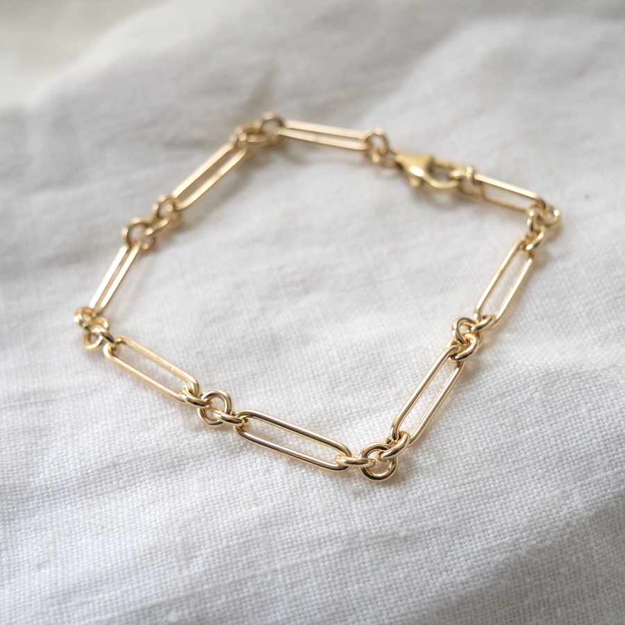 Gold bracelet with mixed link designs, including elongated and rounded links, displayed on a white textured fabric.