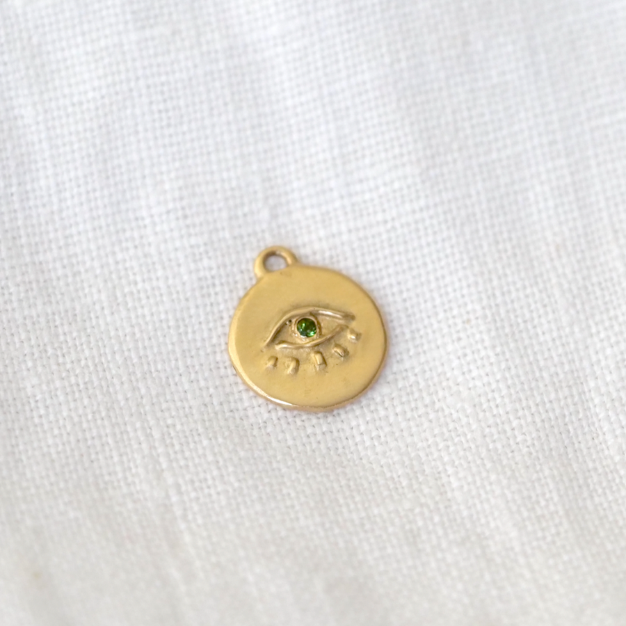 A small, 14k gold charm in the shape of an eye, set with a single green gemstone, resting on a textured white fabric surface showcasing the Seer Totem necklace by Marisa Mason Jewelry.