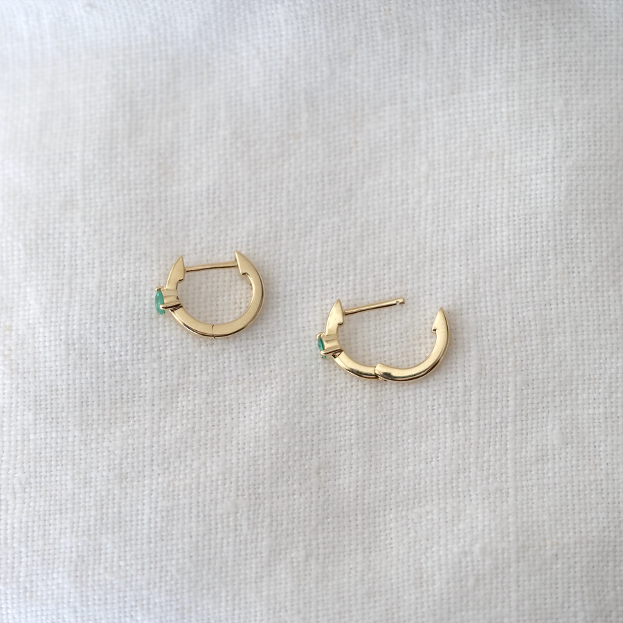 A pair of small White Cloud Co. 3 Prong Wide Emerald Huggie hoop earrings with emerald accents laying on a white linen surface.