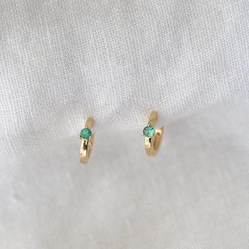 A pair of White Cloud Co. 3 Prong Wide Emerald Huggie earrings with small green gemstones, displayed on a white textured background.