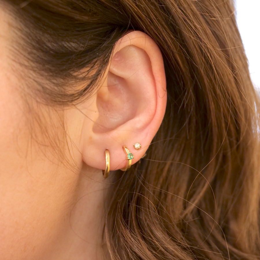 Close-up of a person's ear adorned with three White Cloud Co. 3 Prong Wide Emerald Huggie earrings: an emerald huggie hoop, a stud with a green gem, and a small cuff, set against a