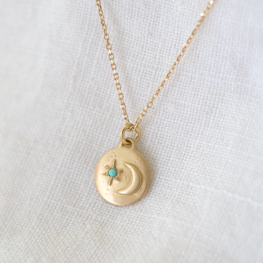 A delicate 14k gold Luna Totem necklace by Marisa Mason Jewelry with a round, textured charm featuring an embossed Crescent Moon and a small turquoise stone, displayed on a white fabric background.