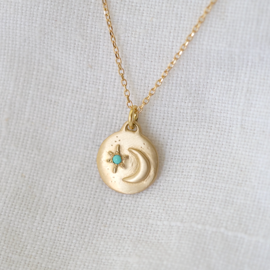 A delicate gold necklace with a Totem pendant from Marisa Mason Jewelry, featuring a small, embedded turquoise gemstone, displayed against a white fabric background.