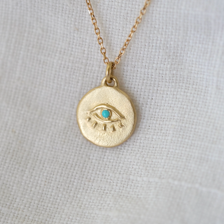 A golden Totem pendant with a turquoise iris, hanging from a delicate chain, set against a textured white fabric background by Marisa Mason Jewelry.