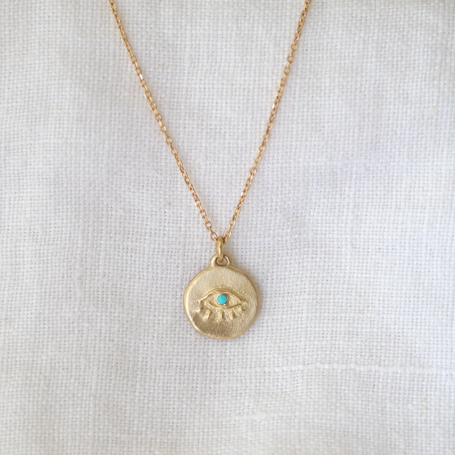 A gold Seer Totem necklace featuring a small turquoise stone in the center, displayed against a white textured background by Marisa Mason Jewelry.