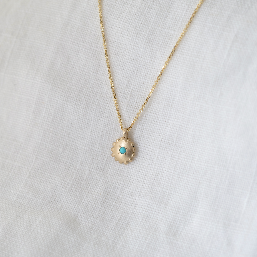 Gold pendant that is round with small granulation around the edge, and a turquoise cabochon at the center. shown on chain with white linen backdrop