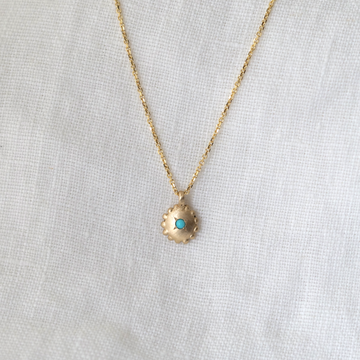Gold pendant that is round with small granulation around the edge, and a turquoise cabochon at the center. shown on chain with white linen backdrop