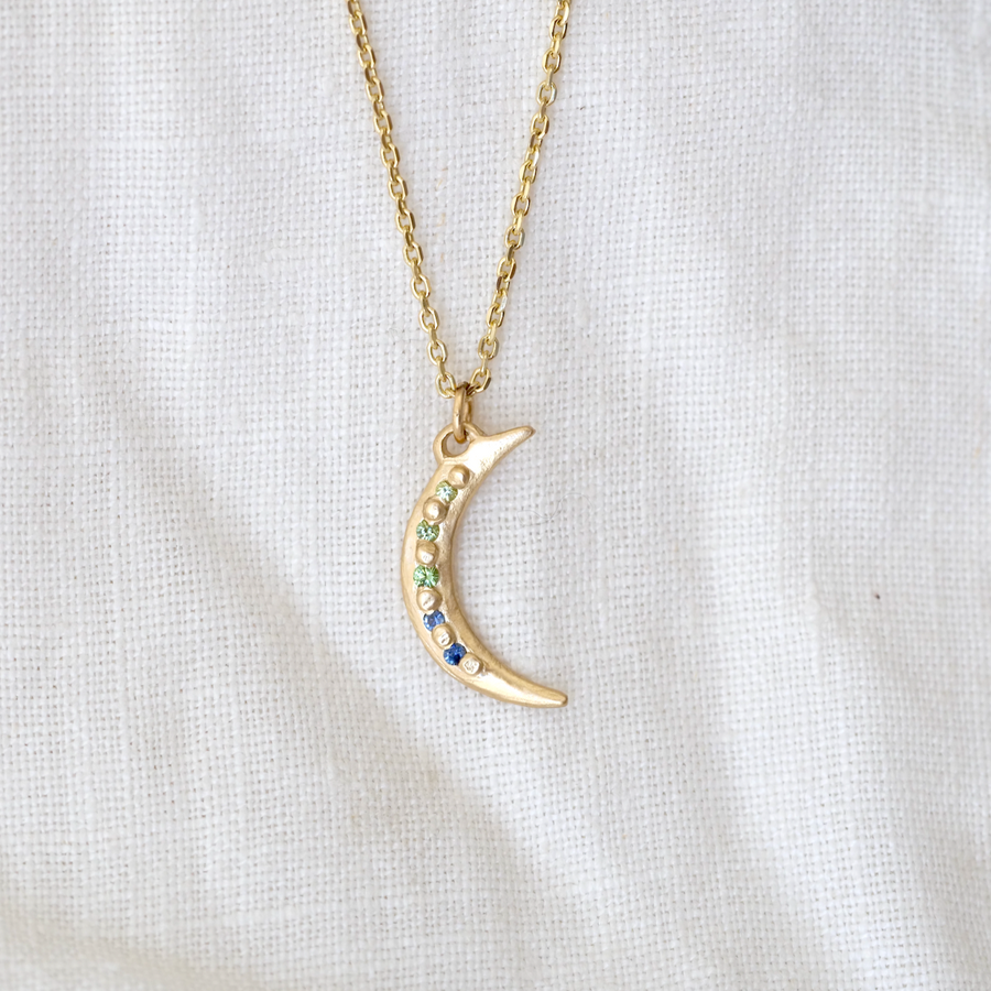 14k crescent moon pendant with green and blue gemstones bead set between gold granulation details down the length of the moon 