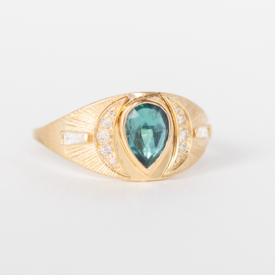 Pear shaped teal tourmaline set at the center of a shield ring with two crescent shapes on either side pave set with small white diamonds, and two diamond parallelograms along the outer edge  with engraved ray lines 