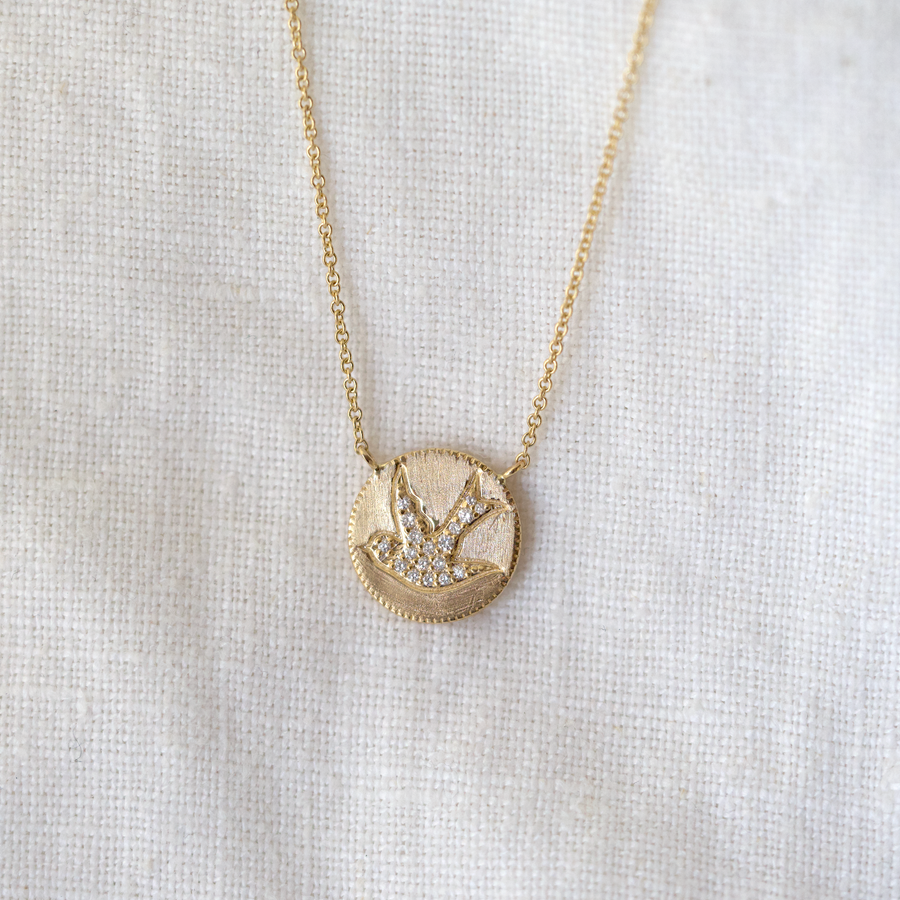 solid 14k pendant features a pave diamond swallow flying across a brushed gold finish, hanging from a 14k chain.