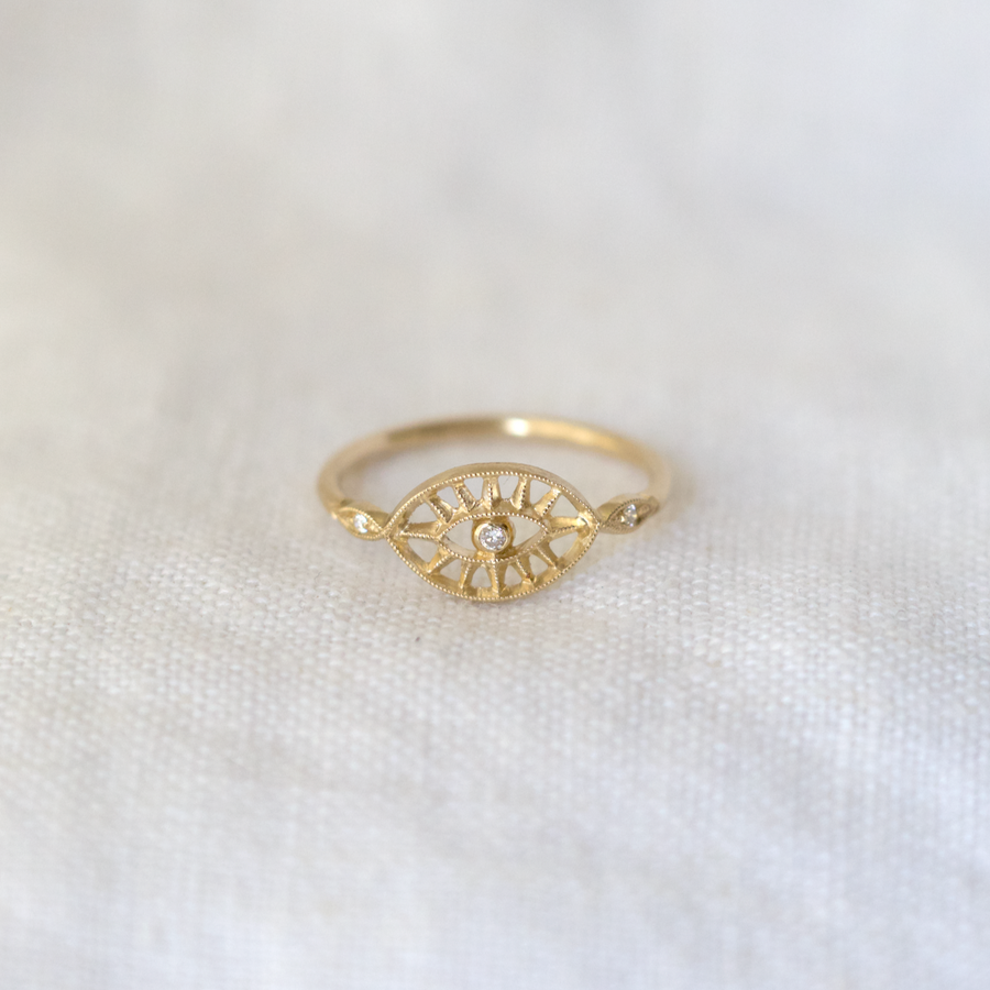 Eye shaped center of ring with diamond center, with negative space cut out making rays around the eye. there are two small diamonds on either side, and a thin gold band