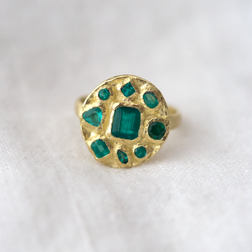 Eight emeralds of varying sizes and shapes, bezel set around a central square cut emerald in the center of a disk of 18k yellow gold. On a simple gold band 