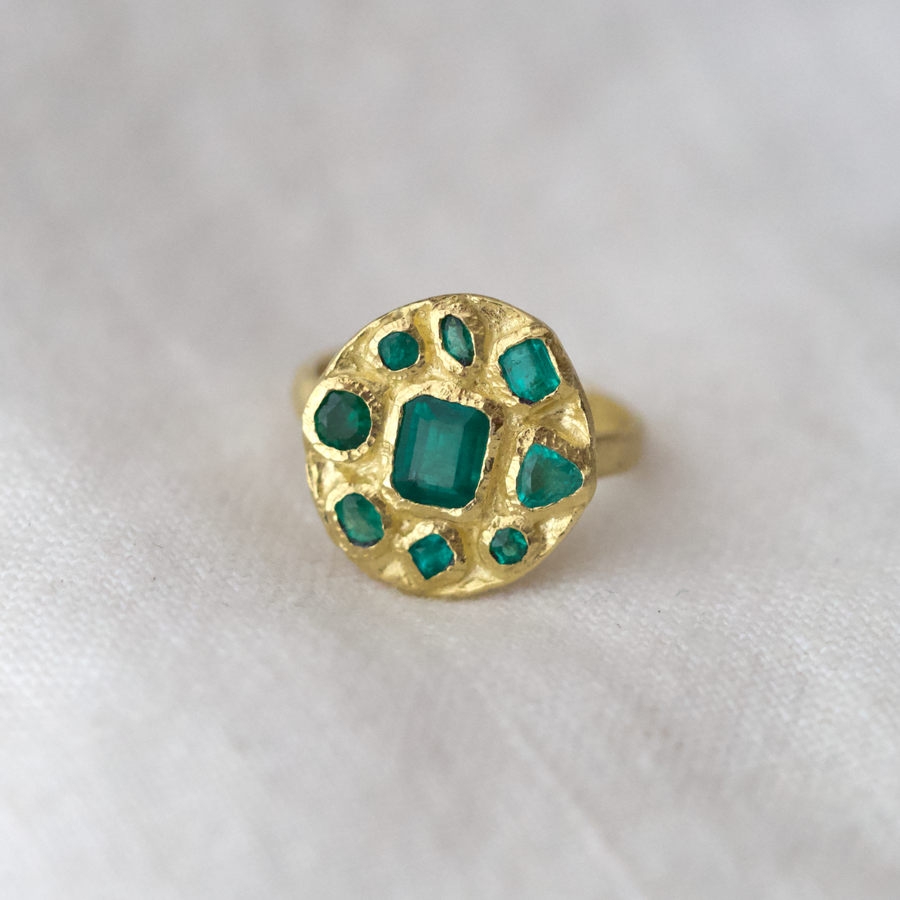 Eight emeralds of varying sizes and shapes, bezel set around a central square cut emerald in the center of a disk of 18k yellow gold. On a simple gold band 