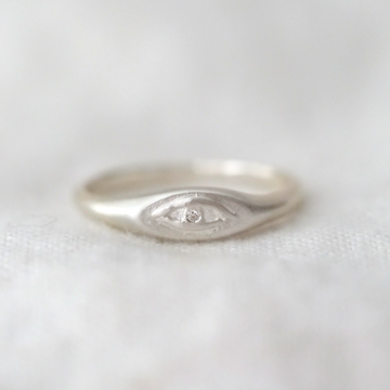 eye is delicately embossed on the front of a thin oval sterling silver signet ring, with a single white diamond in the center. 