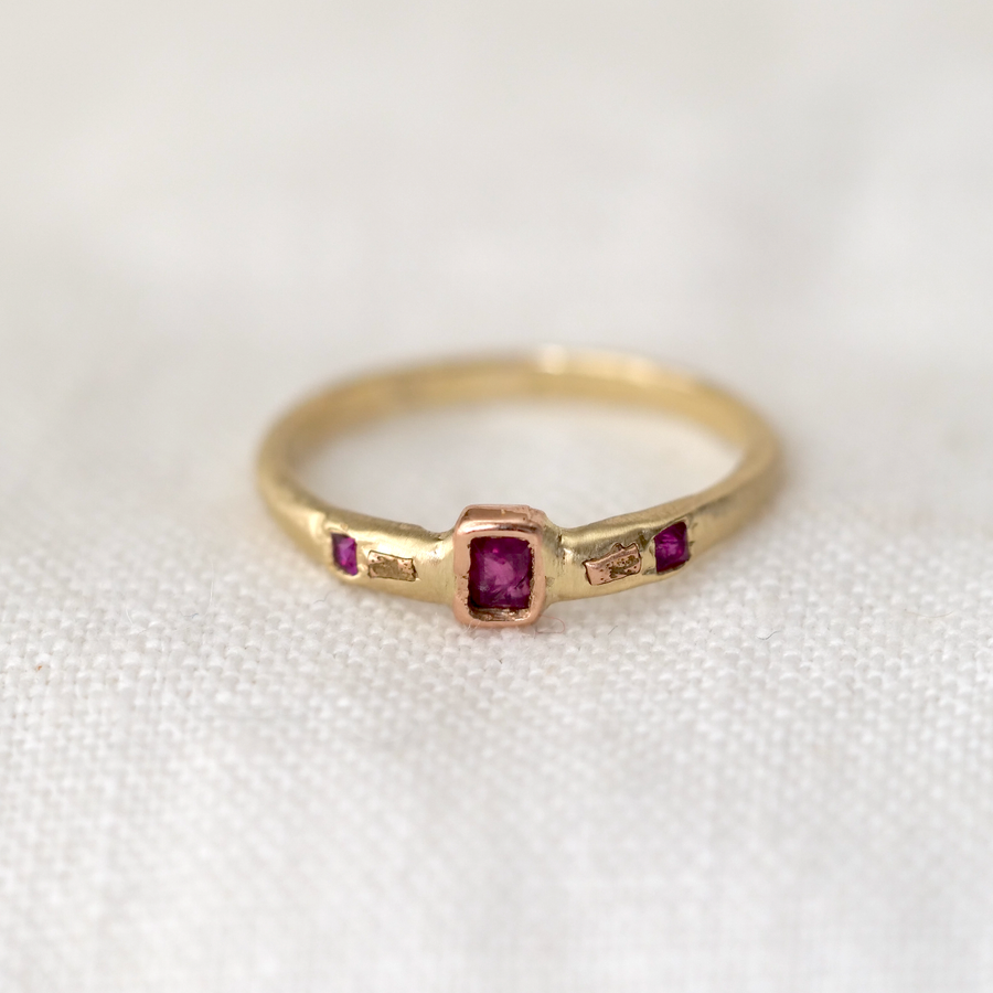 baguette ruby sits perfectly bezel set in 14k Rose Gold on a 14k Yellow Gold band, in between two gold rock sediment specs hand carved in 14k Rose Gold, and two flush set princess cut rubies