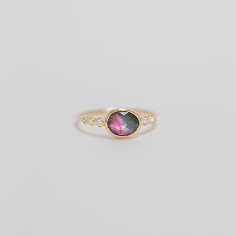 Gold ring with a pink and green gemstone and three diamonds on either side, on a light gray background