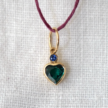Heart-shaped pendant with green gemstone and blue stone on a purple cord against a white background