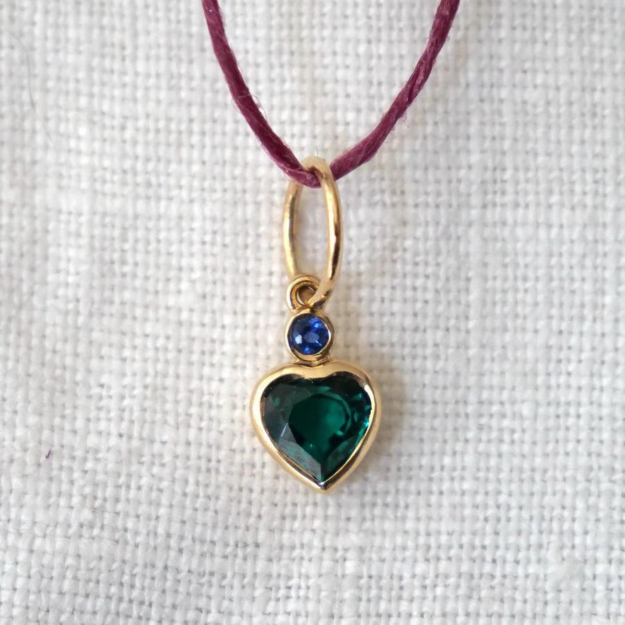 Heart-shaped pendant with green gemstone and blue stone on a purple cord against a white background
