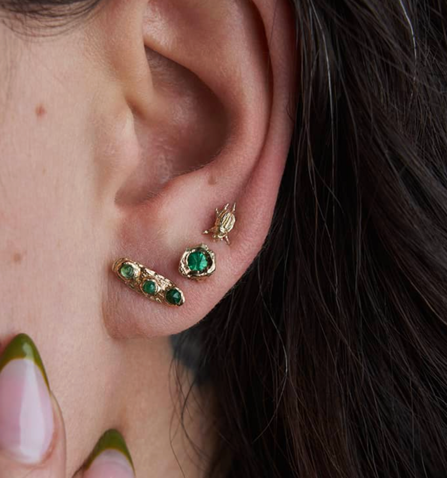 Close-up of an ear wearing gold and green earrings with a blurred background.